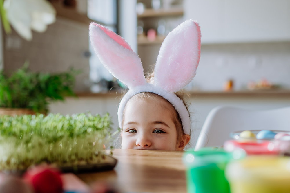 Wielkanocne godziny otwarcia w M1 Radom. Dziewczynka w uszach zająca wielkanocnego, wychylająca się zza stołu.